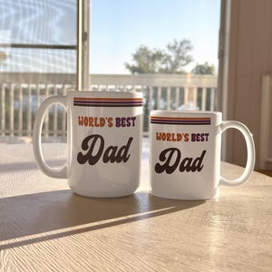 May include: Two white ceramic mugs with the text "WORLD'S BEST Dad" in brown script. The mugs have orange and purple stripes near the top. One mug is larger than the other. The mugs are on a light-colored wooden table.