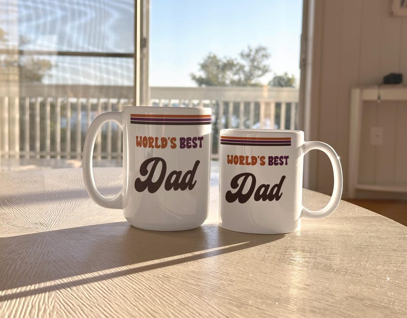 May include: Two white ceramic mugs with the text "WORLD'S BEST Dad" in brown script. The mugs have orange and purple stripes near the top. One mug is larger than the other. The mugs are on a light-colored wooden table.