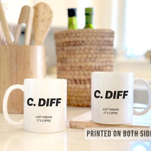 May include: Two white ceramic coffee mugs with the text "C. DIFF" in black, with "JUST KIDDING IT'S COFFEE" below. The mugs are on a wooden cutting board. The phrase "PRINTED ON BOTH SIDES" is at the bottom.