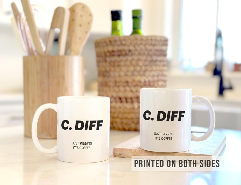 May include: Two white ceramic coffee mugs with the text "C. DIFF" in black, with "JUST KIDDING IT'S COFFEE" below. The mugs are on a wooden cutting board. The phrase "PRINTED ON BOTH SIDES" is at the bottom.