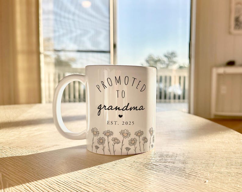 May include: White ceramic mug with the text "PROMOTED TO grandma" in black script, a small heart, and "EST. 2025". The mug features a floral design of daisies. The mug is on a wooden table.
