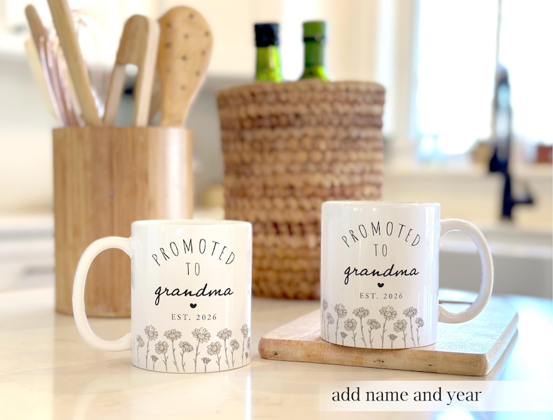 May include: Two white ceramic mugs with the text "PROMOTED TO grandma EST. 2026" in black script. The mugs have a floral design at the bottom. One mug is on a wooden cutting board. The text "add name and year" is at the bottom.