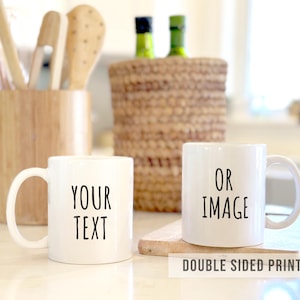 May include: Two white ceramic mugs with black text reading "YOUR TEXT" and "OR IMAGE". The mugs are on a light surface, with kitchen utensils and a woven basket in the background. The text "DOUBLE SIDED PRINT" is at the bottom.