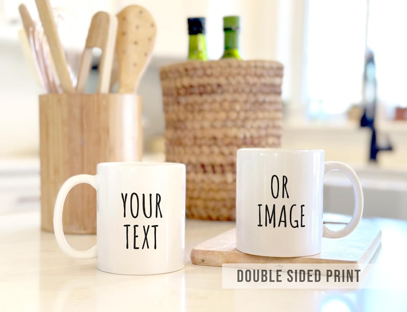 May include: Two white ceramic mugs with black text reading "YOUR TEXT" and "OR IMAGE". The mugs are on a light surface, with kitchen utensils and a woven basket in the background. The text "DOUBLE SIDED PRINT" is at the bottom.