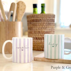 May include: Two ceramic mugs with vertical stripes, one purple and the other green. The purple mug has the name "amelia" and a heart, while the green mug has "lucy" and a heart. The mugs are on a wooden surface.