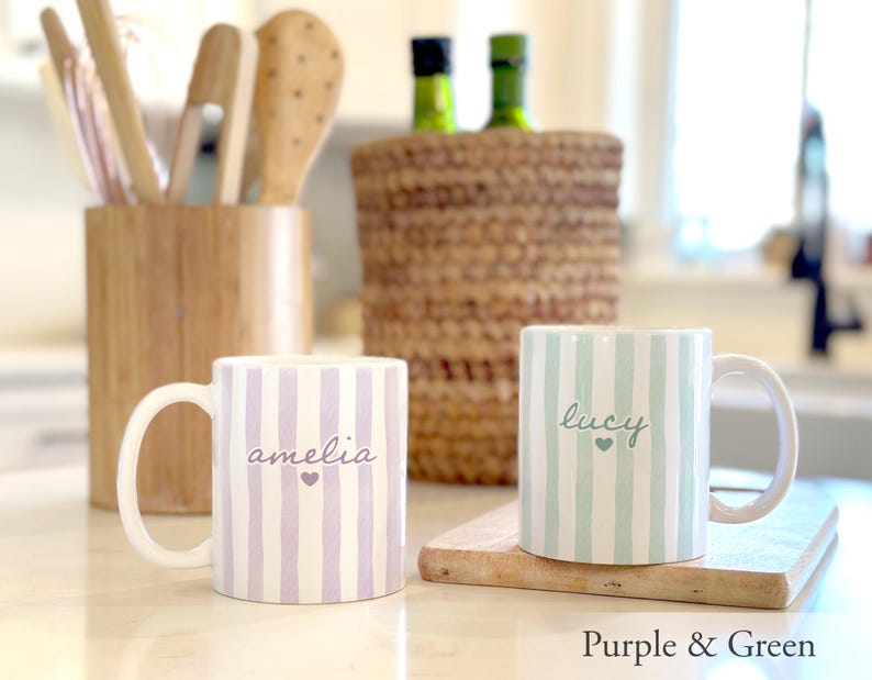 May include: Two ceramic mugs with vertical stripes, one purple and the other green. The purple mug has the name "amelia" and a heart, while the green mug has "lucy" and a heart. The mugs are on a wooden surface.