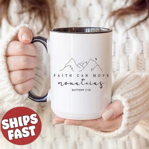 May include: White ceramic mug with a black rim and handle. The mug features a black line drawing of mountains with a sun and moon. The text "FAITH CAN MOVE mountains" is printed in black below the mountains. The text "MATTHEW 17:20" is printed in black below the text.
