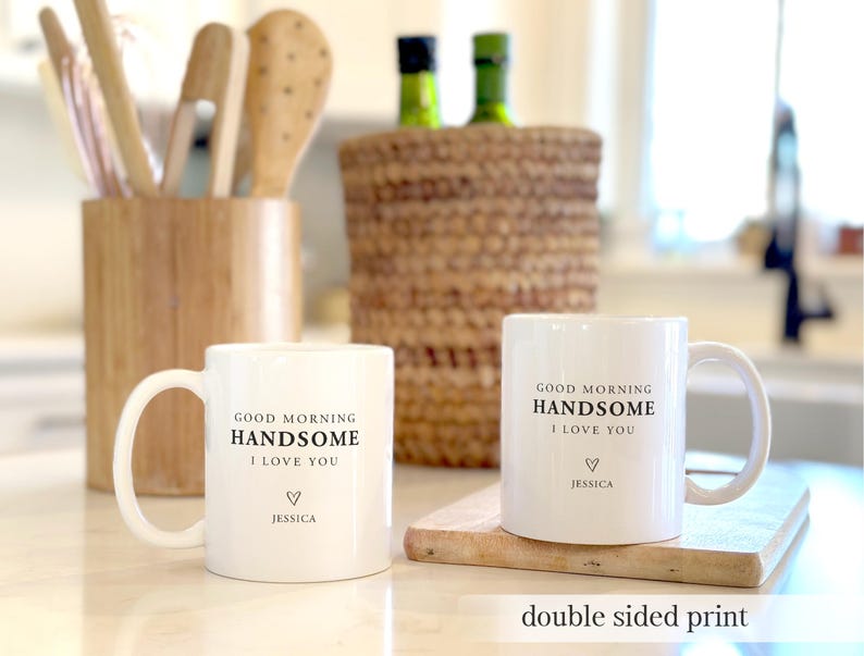 May include: Two white ceramic mugs featuring black printed text that reads GOOD MORNING HANDSOME I LOVE YOU with a small heart symbol and the name JESSICA below it. The mugs have a double-sided print design and a standard handle on the side.