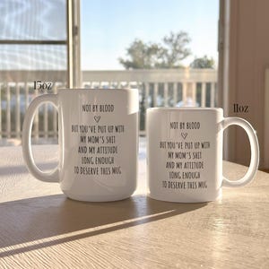 May include: Two white ceramic mugs, 15oz and 11oz, with a curved handle. The mugs have the same black text: "NOT BY BLOOD, BUT YOU'VE PUT UP WITH MY MOM'S SHIT AND MY ATTITUDE LONG ENOUGH TO DESERVE THIS MUG."