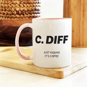 May include: White ceramic coffee mug with a pink interior and handle. The mug features the text "C. DIFF" in bold black letters, with the phrase "JUST KIDDING IT'S COFFEE" below. The mug sits on a wooden tray.