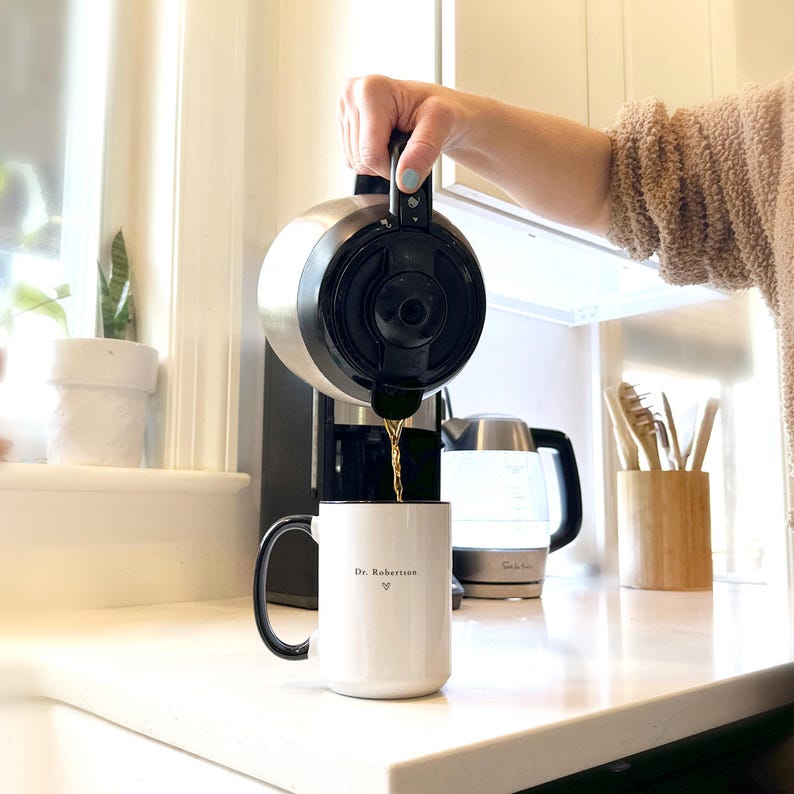 Pu&ograve; includere: Una caffettiera in acciaio inossidabile versa il caff&egrave; in una tazza bianca con manico nero. La tazza ha la scritta "Dr. Robertson" e un simbolo a cuore. Un bollitore e un portautensili in legno sono sullo sfondo.