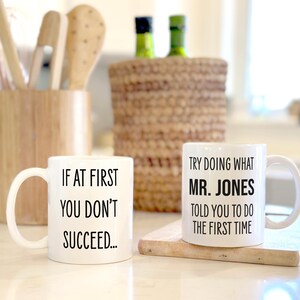 May include: Two white ceramic mugs with black text. One mug reads "IF AT FIRST YOU DON'T SUCCEED..." and the other reads "TRY DOING WHAT MR. JONES TOLD YOU TO DO THE FIRST TIME". The mugs are on a light-colored surface.