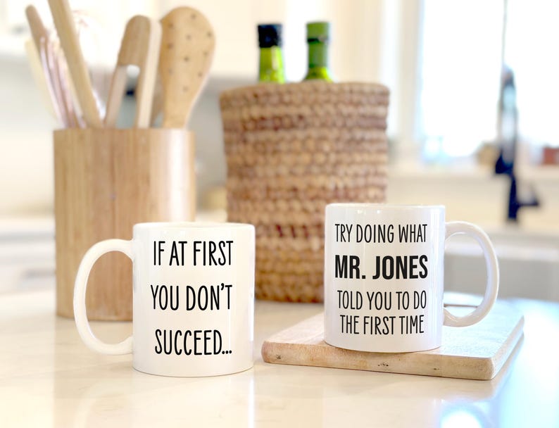 May include: Two white ceramic mugs with black text. One mug reads "IF AT FIRST YOU DON'T SUCCEED..." and the other reads "TRY DOING WHAT MR. JONES TOLD YOU TO DO THE FIRST TIME". The mugs are on a light-colored surface.