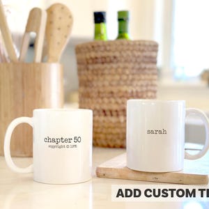 May include: Two white ceramic mugs with black text. One mug says "chapter 50 copyright 1975", the other says "sarah". The mugs are on a light-colored surface, with wooden utensils and a woven basket in the background.