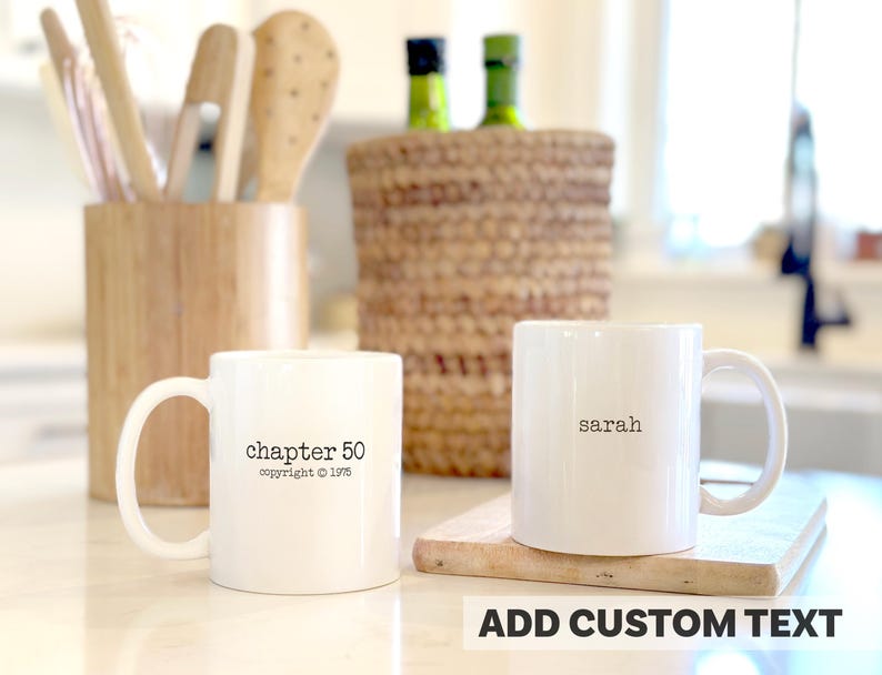 May include: Two white ceramic mugs with black text. One mug says "chapter 50 copyright 1975", the other says "sarah". The mugs are on a light-colored surface, with wooden utensils and a woven basket in the background.