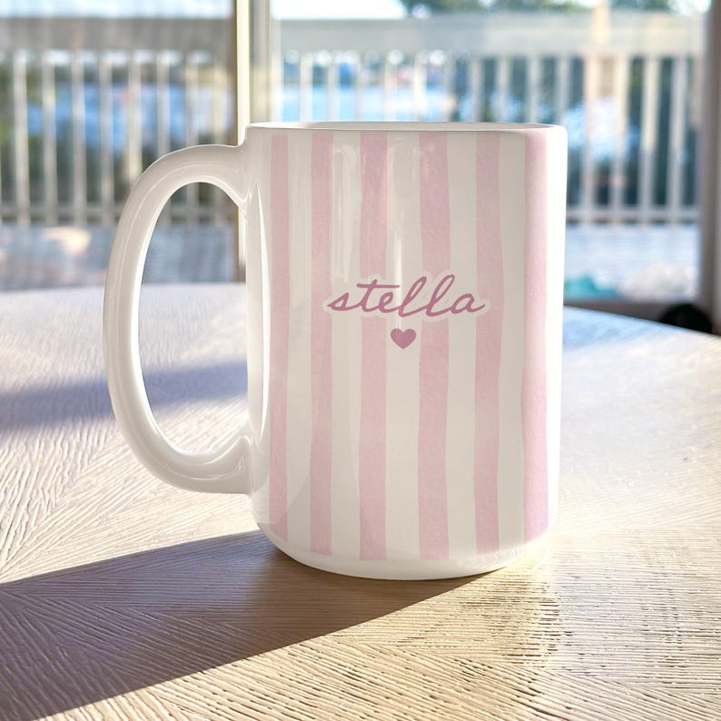 May include: White ceramic mug with vertical pink stripes. The word "stella" is written in a cursive font, with a small pink heart below the name. The mug has a large handle.