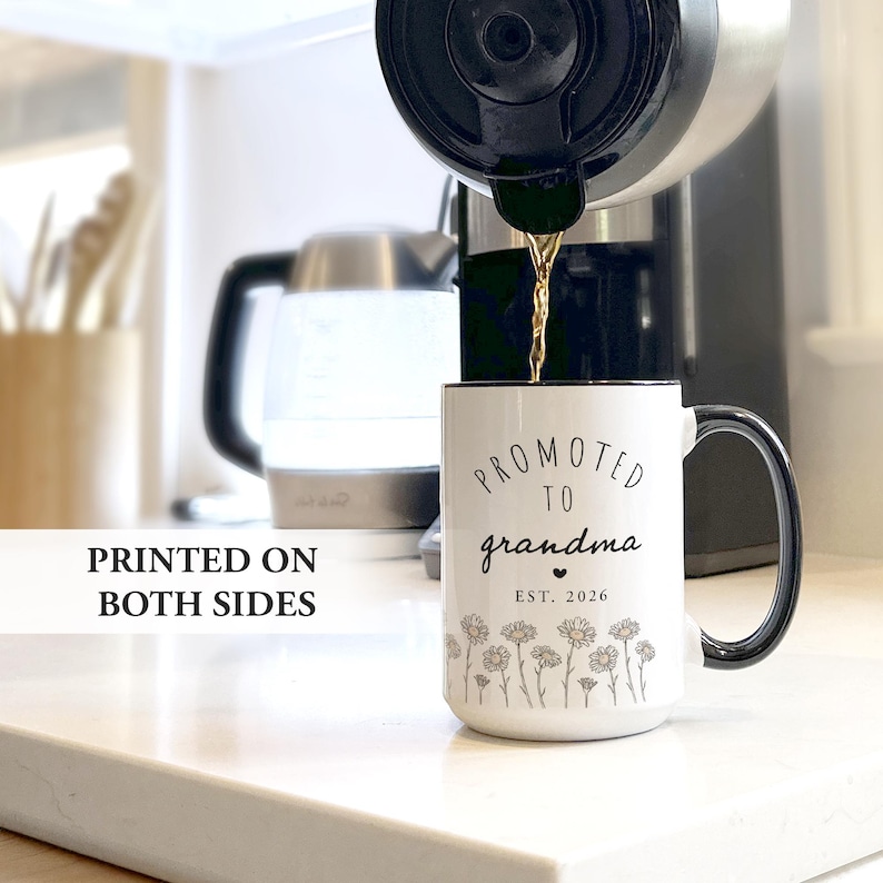 May include: White ceramic mug with a black handle and rim, featuring the text "Promoted to grandma" and "EST. 2026". The mug is decorated with a floral design and the words "Printed on both sides". Coffee is being poured into the mug.