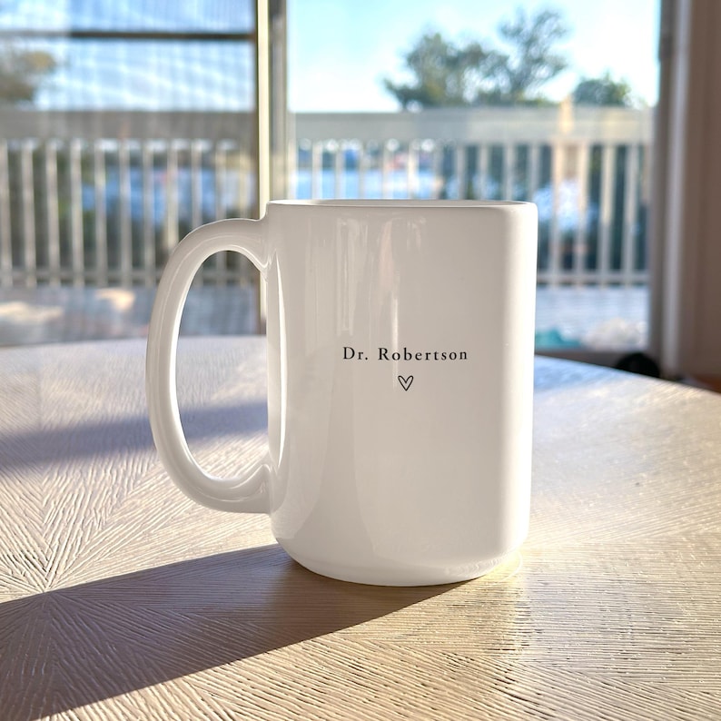 Pu&ograve; includere: Una tazza di ceramica bianca con un manico grande. La tazza presenta la scritta "Dr. Robertson" e un simbolo a forma di cuore. La tazza &egrave; appoggiata su una superficie chiara.