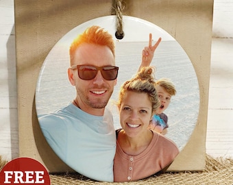 May include: A circular ceramic ornament featuring a printed photograph of two adults and a child. The adult on the left wears dark sunglasses and a light blue shirt. The adult on the right wears a peach-colored top. A child is visible behind them.