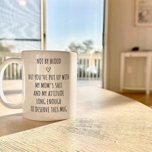 May include: White ceramic mug with a large handle, featuring black text that reads "NOT BY BLOOD, BUT YOU'VE PUT UP WITH MY MOM'S SHIT AND MY ATTITUDE LONG ENOUGH TO DESERVE THIS MUG." The mug is on a light-colored wooden table.