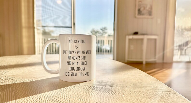 May include: White ceramic mug with a large handle, featuring black text that reads "NOT BY BLOOD, BUT YOU'VE PUT UP WITH MY MOM'S SHIT AND MY ATTITUDE LONG ENOUGH TO DESERVE THIS MUG." The mug is on a light-colored wooden table.