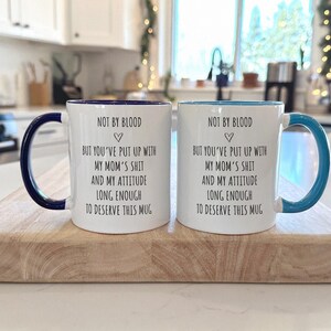 May include: Two white ceramic mugs with dark blue and light blue handles and rims. Each mug has the text "NOT BY BLOOD" above a heart symbol, followed by a humorous message. The mugs are displayed on a wooden cutting board.