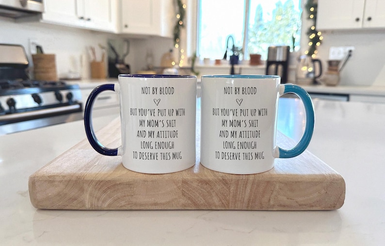 May include: Two white ceramic mugs with dark blue and light blue handles and rims. Each mug has the text "NOT BY BLOOD" above a heart symbol, followed by a humorous message. The mugs are displayed on a wooden cutting board.
