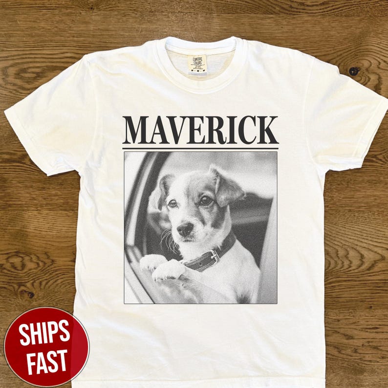 May include: White t-shirt with the word "MAVERICK" printed in black above a black and white image of a dog looking out of a car window. The dog is wearing a collar.