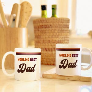 May include: Two white ceramic mugs with the text "WORLD'S BEST Dad" in a retro font. Each mug has a brown and red stripe design. The mugs are on a wooden cutting board.
