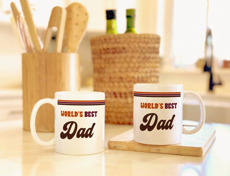 May include: Two white ceramic mugs with the text "WORLD'S BEST Dad" in a retro font. Each mug has a brown and red stripe design. The mugs are on a wooden cutting board.