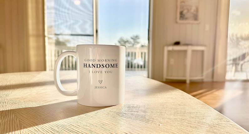 May include: A white ceramic mug featuring the black printed text GOOD MORNING HANDSOME I LOVE YOU, a small heart symbol, and the name JESSICA centred below. The mug has a standard handle and sits on a light-coloured wooden surface in bright light.