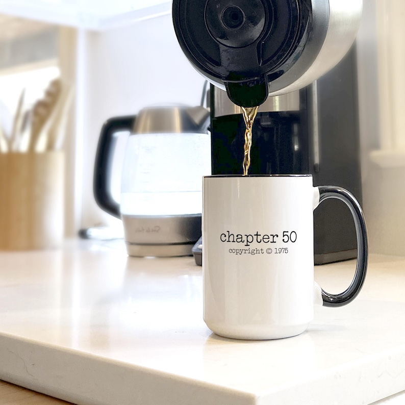 May include: A white coffee mug with a black rim and handle is being filled with coffee. The mug has the text "chapter 50 copyright &copy; 1975" printed on it. A coffee maker and a kettle are in the background.