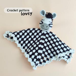 Pode incluir: Um amor de crochê feito à mão com uma cabeça de zebra e um cobertor xadrez preto e branco. A zebra tem orelhas cinzentas, um focinho cinzento e listras pretas. O cobertor tem uma borda branca com vieiras. O texto "Crochet pattern lovey" está no canto superior esquerdo.
