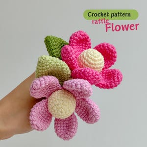 May include: Crocheted flower rattle in shades of pink, yellow, and green. The image includes the text "Crochet pattern rattle Flower". The handmade rattle features a flower design with soft, textured petals and a central sphere.