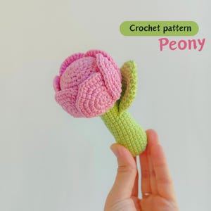 Op de afbeelding: Een handgemaakte gehaakte pioenroos in roze en groen. De bloemkop is helderroze, met een strak gewikkelde knop. De stengel en bladeren zijn lichtgroen. Tekst op de afbeelding luidt "Crochet pattern Peony."