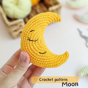以下が含まれることがあります： 手作りの、笑顔の顔をした黄色の編み物で作られた三日月が手に持たれています。 三日月は、質感のある糸でできており、黒い刺繍の目と口があります。 画像には「Crochet pattern Moon」というテキストが表示されています。