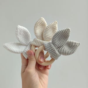 May include: Three crochet baby teethers in white, cream, and gray. Each teether features a wooden ring and a bunny ear design. The teethers are handmade and suitable for infants.