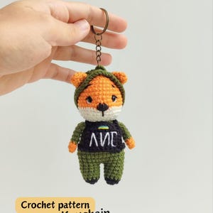 May include: A crocheted fox keyring in orange, white, and green. The fox wears a dark blue top with white text and a green hat. It is attached to a metal ring. The text "Crochet pattern Keychain" is in a yellow box.