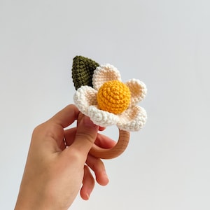 May include: A handmade crochet baby rattle in the shape of a flower. The flower has white petals, a yellow center, and a green leaf. The rattle has a wooden ring for easy gripping.
