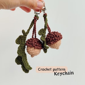 May include: Crocheted acorn keychain with brown caps and tan bottoms, attached to a silver clasp. Green crocheted leaves and stems are also included. The text "Crochet pattern Keychain" is visible.