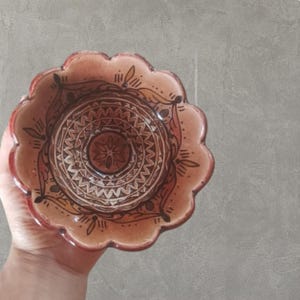 Puede incluir: Un pequeño cuenco decorativo de cerámica con borde festoneado. El cuenco presenta una combinación de colores marrón y beige con intrincados patrones geométricos pintados a mano. El borde es de un tono marrón más oscuro.