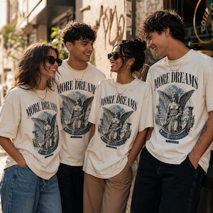 May include: Off-white short-sleeve t-shirts featuring a graphic print of an angel with wings, the text "CHASE MORE DREAMS" in a curved font, and the smaller text "WORK FOR IT" at the bottom. The design includes a circular frame and pillars.