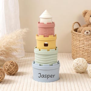 May include: A stackable tower toy in pastel colors, resembling a castle. The tower has a white top, pink, yellow, and green sections, and a blue base with the name "Jasper". Decorative balls and teddy bears are in the background.