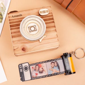 May include: A wooden camera-shaped box with the name "Amanda" engraved on it, and a decorative lens. A film roll keychain with photos is next to the box. The film roll is black and yellow.