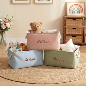 Peut inclure: Trois bacs de rangement rectangulaires en tissu aux couleurs pastel. Le bac rose porte un arc-en-ciel et le nom "Veirty". Le bleu a un voilier et le nom "Apollo". Le vert a une empreinte de patte et le nom "Cyrus". Les bacs sont remplis de jouets et ont des poignées en corde.