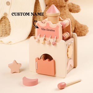 Peut inclure: Un château de jouet en bois aux couleurs pastel, avec une tourelle rose et divers éléments interactifs. Le château porte le nom "Ellie Christine" et comprend une étoile, un croissant et un maillet.