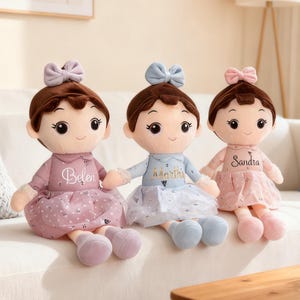 May include: Three plush dolls with brown hair, bows, and dresses in pink and blue hues. Each doll has a name embroidered on the dress: Belen, Martha, and Sandra. The dolls are sitting on a white couch.