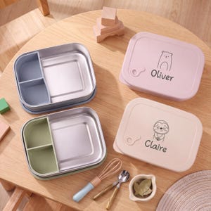 Può includere: Due scatole bento rettangolari in acciaio inossidabile con scomparti divisi. Un coperchio è rosa con l'illustrazione di un orso e il nome Oliver, l'altro è beige con l'illustrazione di un bradipo e il nome Claire. Include una frusta, un cucchiaio e una piccola ciotola.