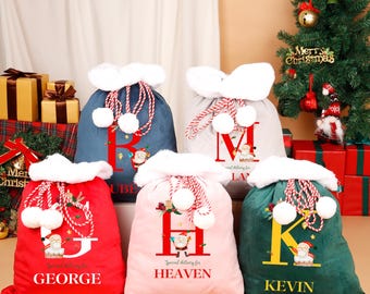 May include: A selection of Christmas gift bags in shades of red, pink, blue, grey, and green. Each bag has a white fluffy trim, a large initial, and a Santa Claus graphic. The names "GEORGE", "HEAVEN", "KEVIN", "RUBEN", and "LA" are printed on the bags.