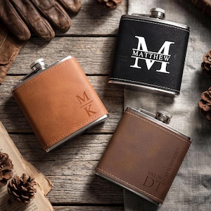 Op de afbeelding: Drie gepersonaliseerde heupflessen met roestvrijstalen doppen. Eén is zwart met een witte initiaal en naam, één is bruin met initialen en de derde is bruin met initialen en het woord "Groomsman".